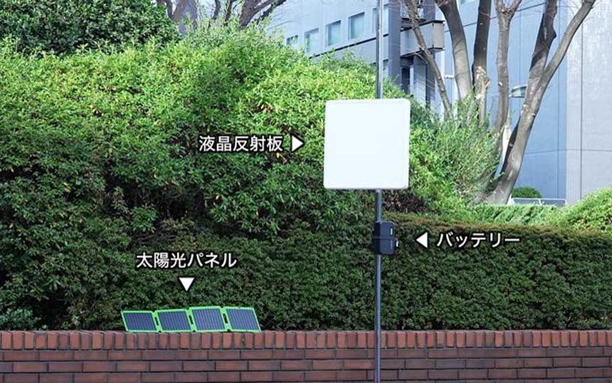 屋外に設置された液晶反射板。液晶反射板は50センチ四方、重量8キロで、スマートフォンでON/OFF、散乱パターンの変更が可能（ビル街の植え込みの前に白い液晶反射板とバッテリーがポールに取り付けられている。小型の太陽光パネルが植え込みのレンガ枠に置かれている）