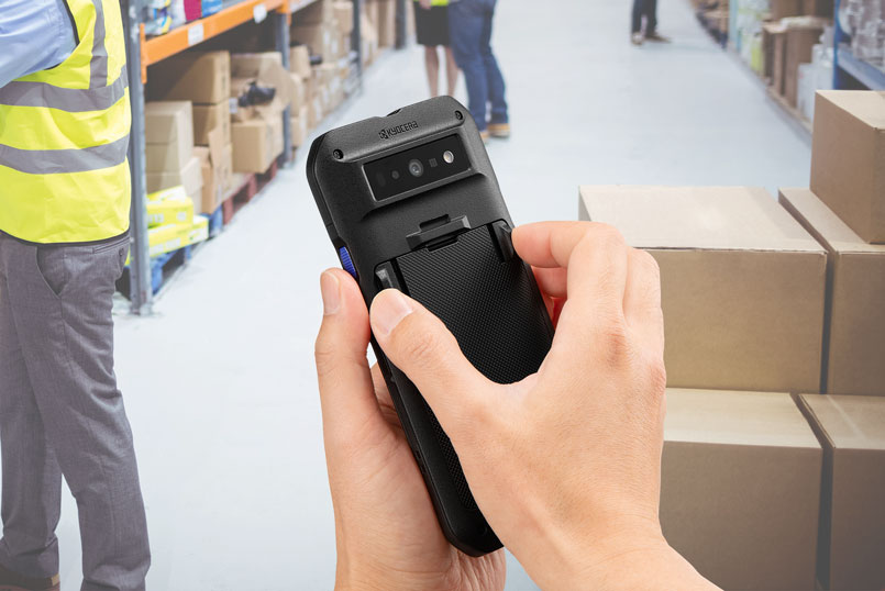 ホットスワップのイメージ。倉庫内の通路で、作業者が京セラ製スマートフォン「DuraForce EX2」の背面カバーを外し、バッテリーを交換している様子。周囲には棚と段ボール箱が並び、現場作業中でも電源を切らずにバッテリー交換できるホットスワップ機能を示している。