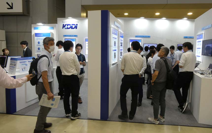 Group of professionals at a trade show booth with screens and posters, KDDI branding visible in the background.