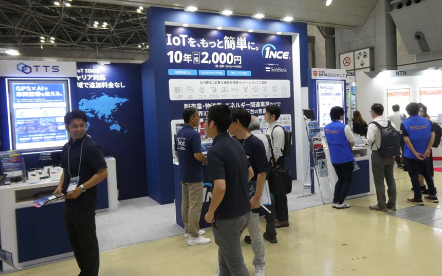 Trade show booth with blue panels and attendees, promoting IoT services at a tech expo panel display in Japanese signage in the background.