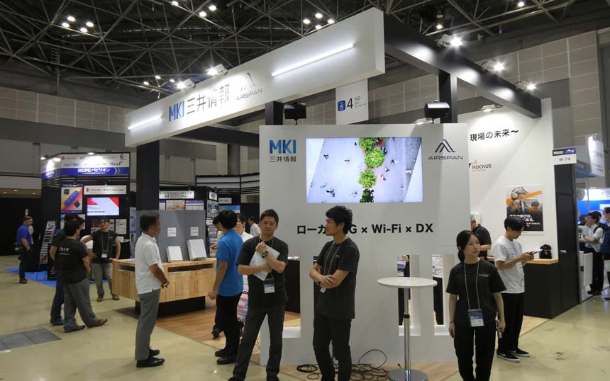 Trade show booth with MKI branding, large white overhead structure, a wall-mounted screen showing greenery, and several attendees standing around.