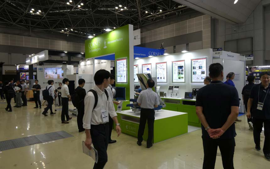 Exhibition hall scene with a bright lime-green trade show booth, display screens, and attendees walking around and engaging with exhibits.