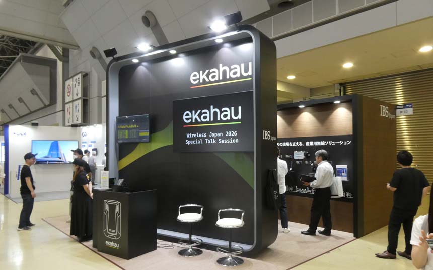 Ekahau trade show booth with a curved black wall, logo, a mounted screen showing 'Wireless Japan 2026 Special Talk Session' and two white stools in front of it