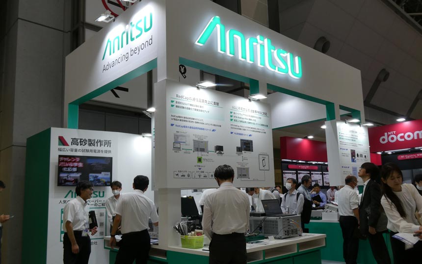 Anritsu exhibition booth at a trade show with teal-and-white display, large Anritsu logo, and technical posters as staff assist attendees.