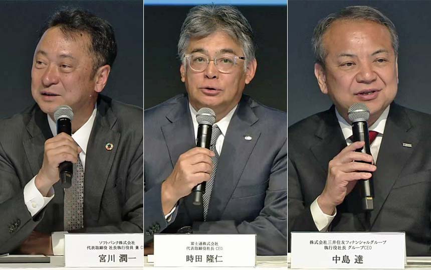 Three men in suits seated at a panel, each holding a handheld microphone and speaking during a conference.