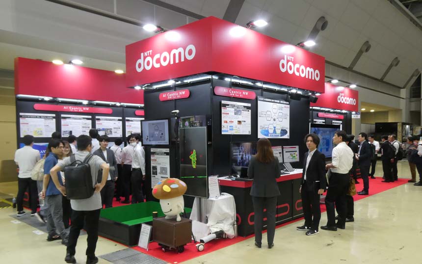 NTT Docomo exhibition booth at a tech expo with staff, screens, and a mascot figure on display hostly interacting visitors nearby