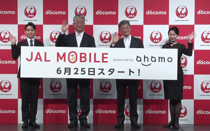 Four people in business attire hold a banner announcing JAL MOBILE powered by ahamo, while waving, in front of a red-and-white checkered backdrop with logos.