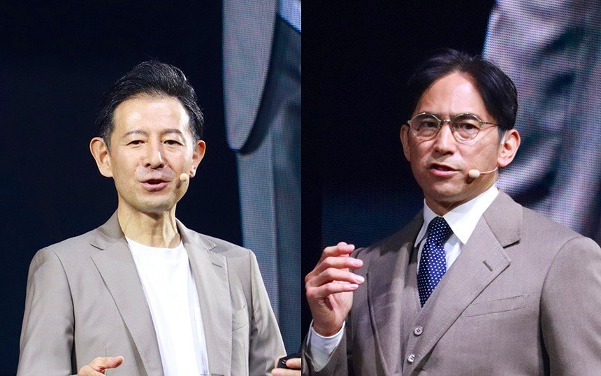Two men in light gray suits on a stage, each wearing a headset microphone, speaking to an audience on a dark backdrop.