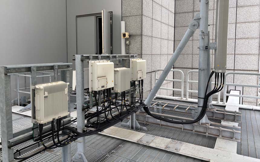 Outdoor rooftop with a row of white electrical boxes mounted on a metal frame, multiple black cables running between them and a tall utility pole.