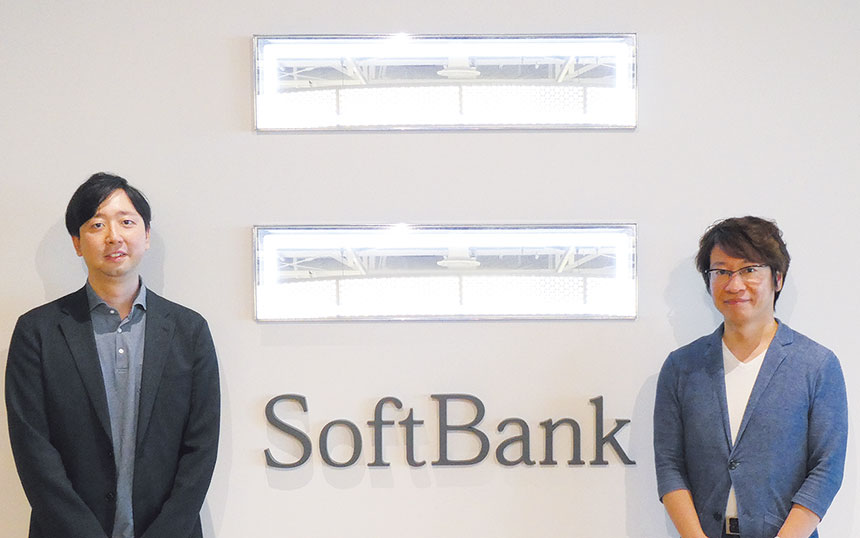Two men in blazer jackets stand on either side of a SoftBank logo in a bright office lobby.