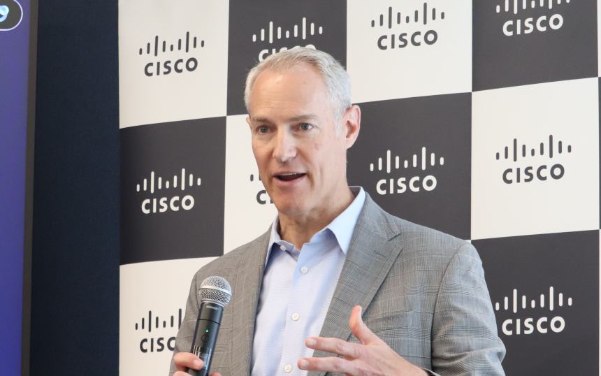 Man in a light blue shirt and gray blazer speaks into a handheld microphone at a Cisco event, with Cisco logos on the backdrop behind him.